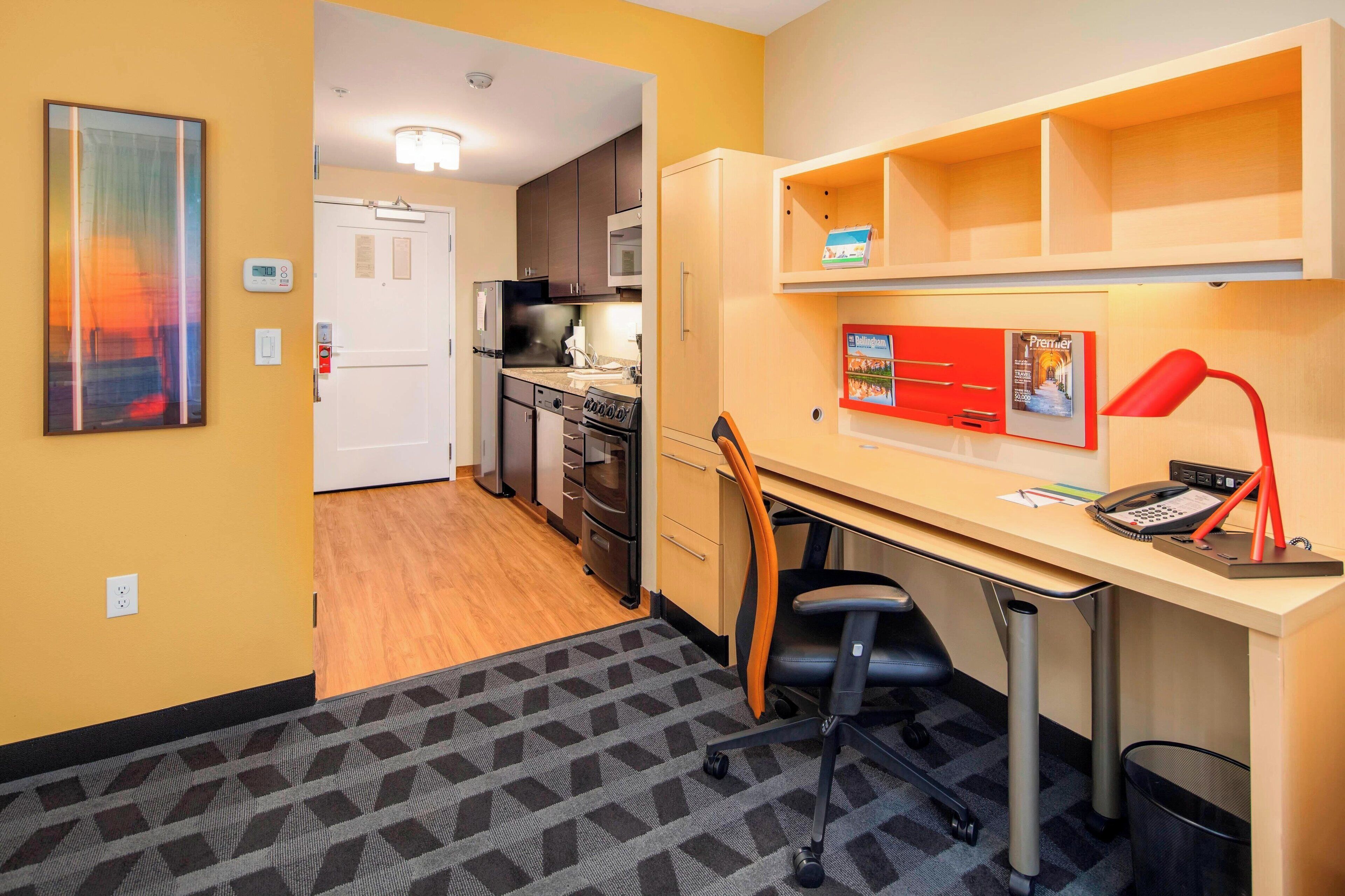 Une chambre d’hôtel compacte comprenant un coin cuisine, un bureau avec une chaise, une lampe rouge et une affiche encadrée au mur.
