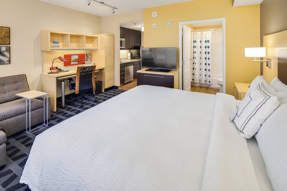 A hotel room with a large bed, a desk with a computer, a TV, and a kitchenette.
