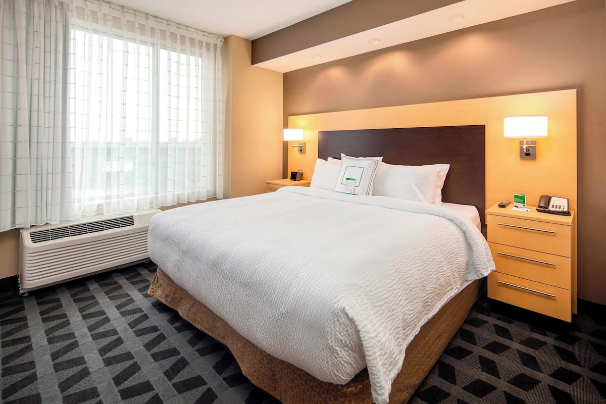 A hotel room with a large bed, a bedside table with a lamp, a heater, and a window with curtains.
