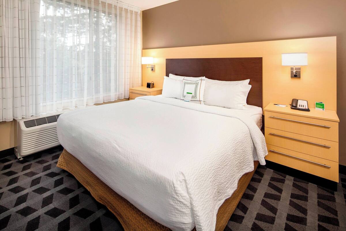A hotel room with a large bed, two bedside tables, a heater, and a window with sheer curtains.