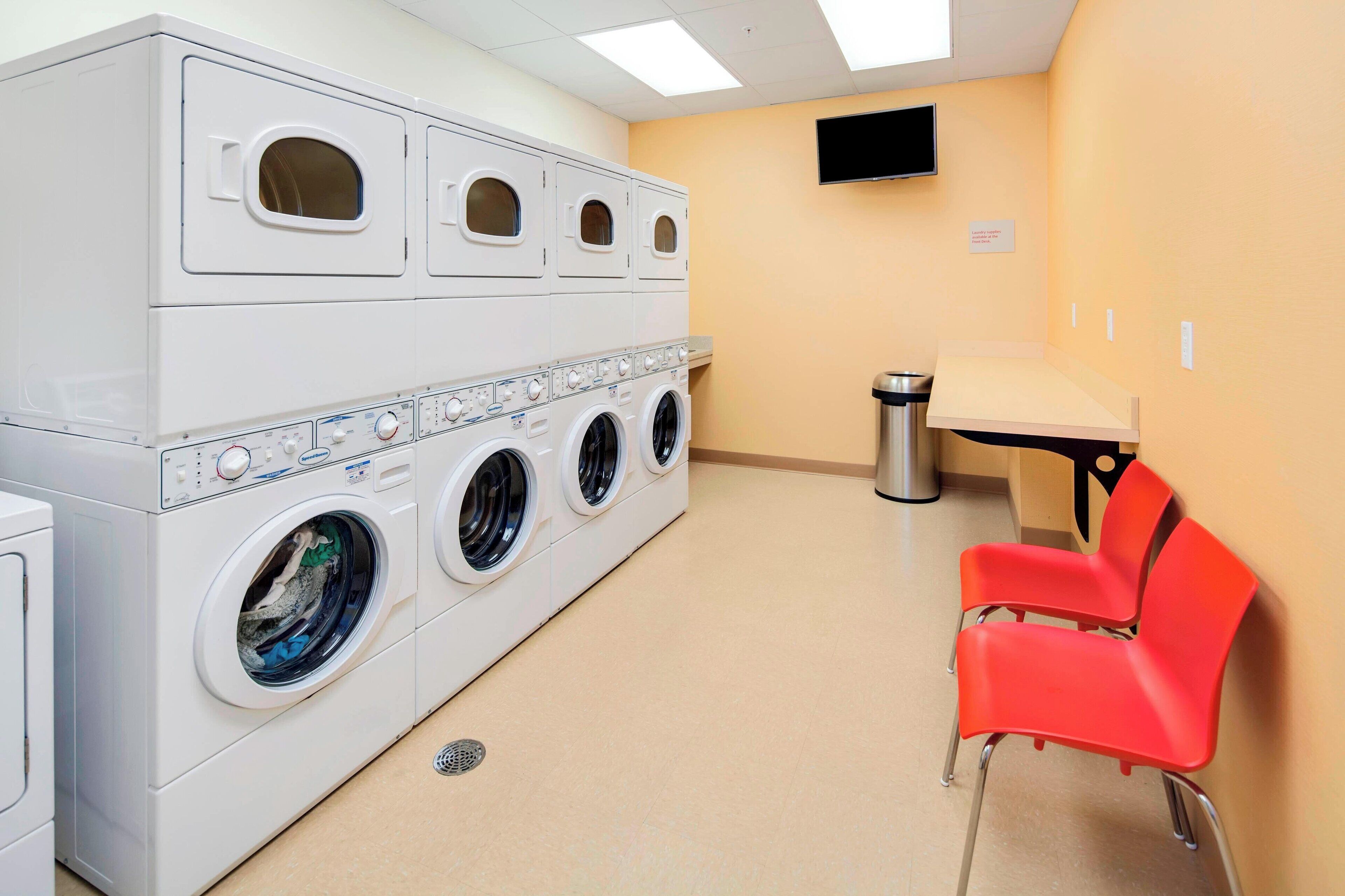 A laundry room with multiple washing machines, a trash bin, and a table with two red chairs.