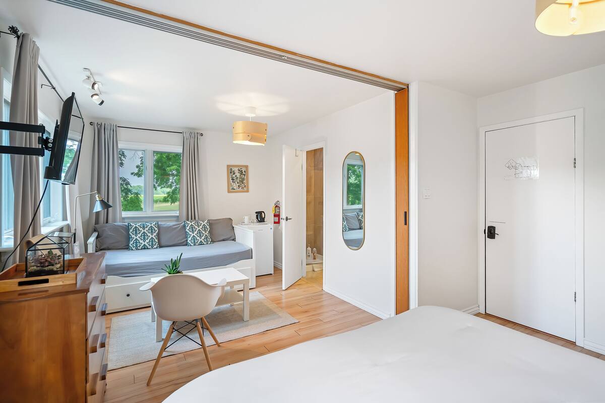 A modern bedroom with a bed, bedside table, chair, and a desk with a lamp. There is a window with curtains, a door, and a view of greenery outside.