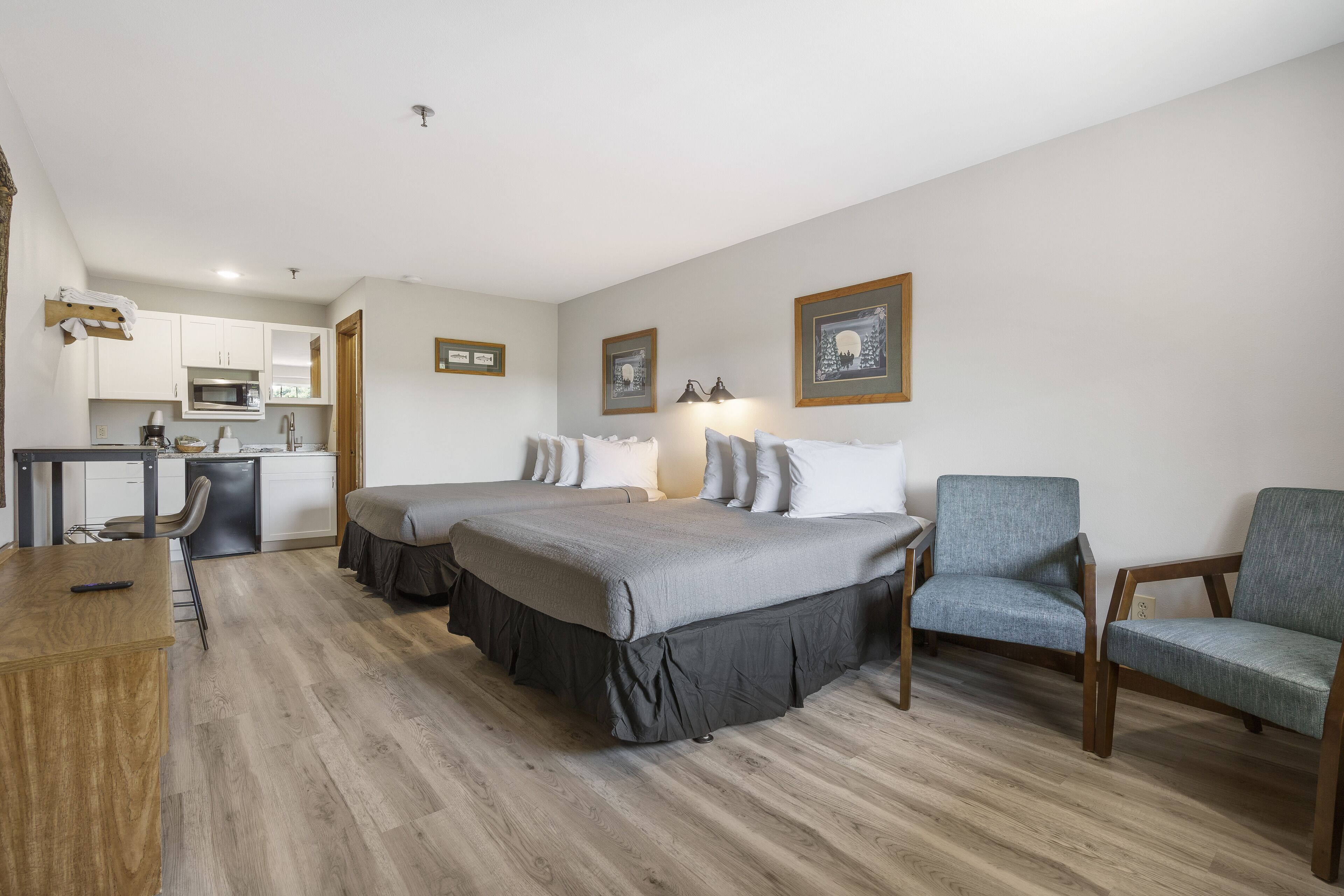 A hotel room with a large bed, two bedside tables, a desk, a chair, a kitchenette, and framed pictures on the wall.