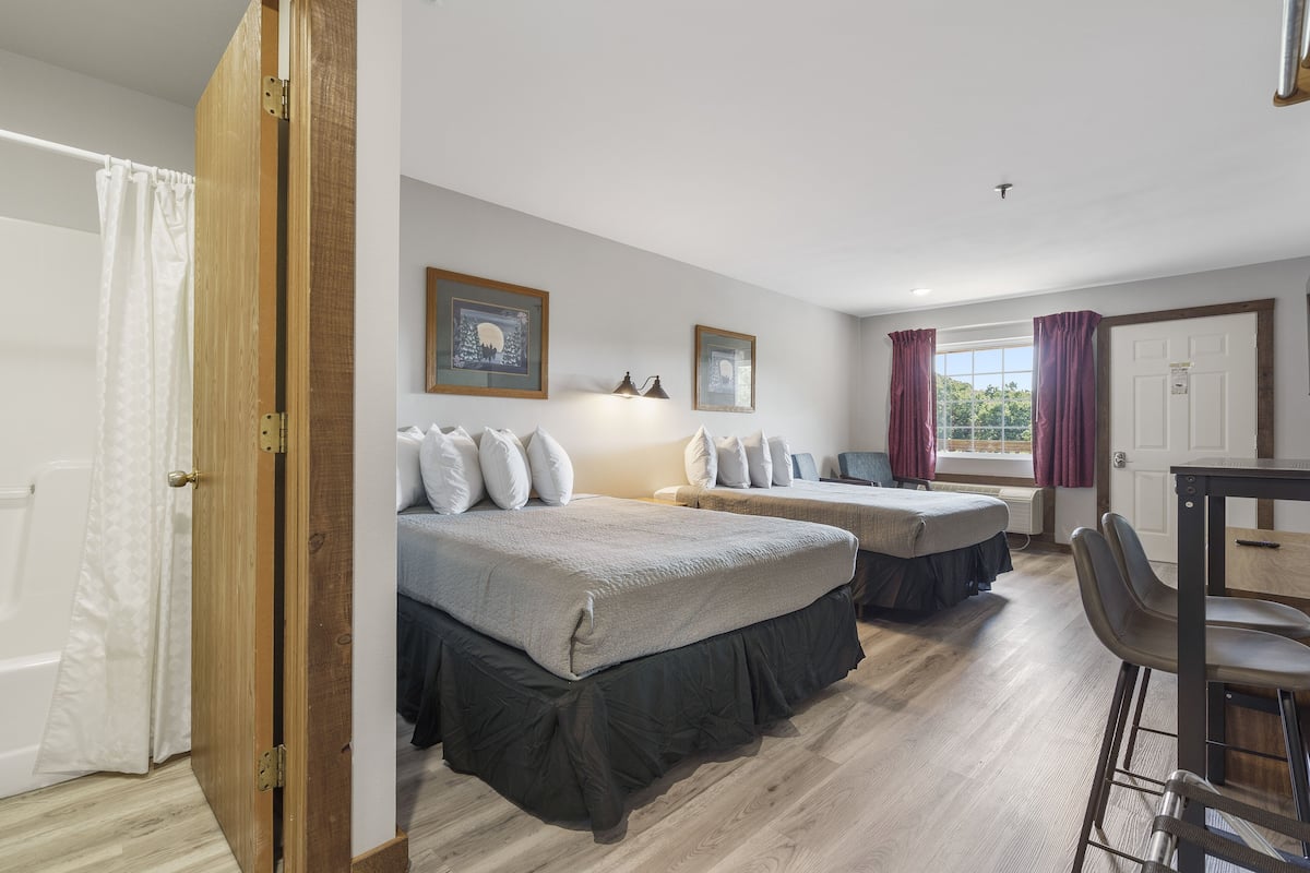 Une pièce avec deux lits, une table et une chaise. Il y a une fenêtre avec des rideaux, une porte et un mur orné de tableaux.