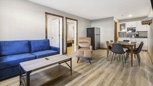 Sala de estar com um sofá azul, uma mesa de centro de madeira e uma poltrona bege. Há também um espaço de jantar com mesa e cadeiras, e uma cozinha com armários e eletrodomésticos brancos.