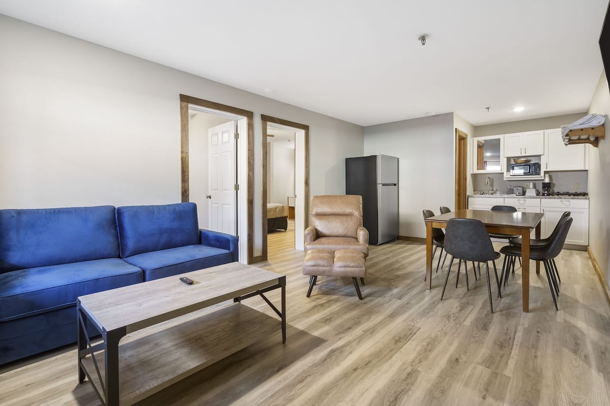 Un salon avec un canapé bleu, une table basse en bois et un fauteuil beige. On y trouve également un coin repas avec une table et des chaises, ainsi qu’une cuisine dotée de meubles blancs et d’appareils électroménagers.