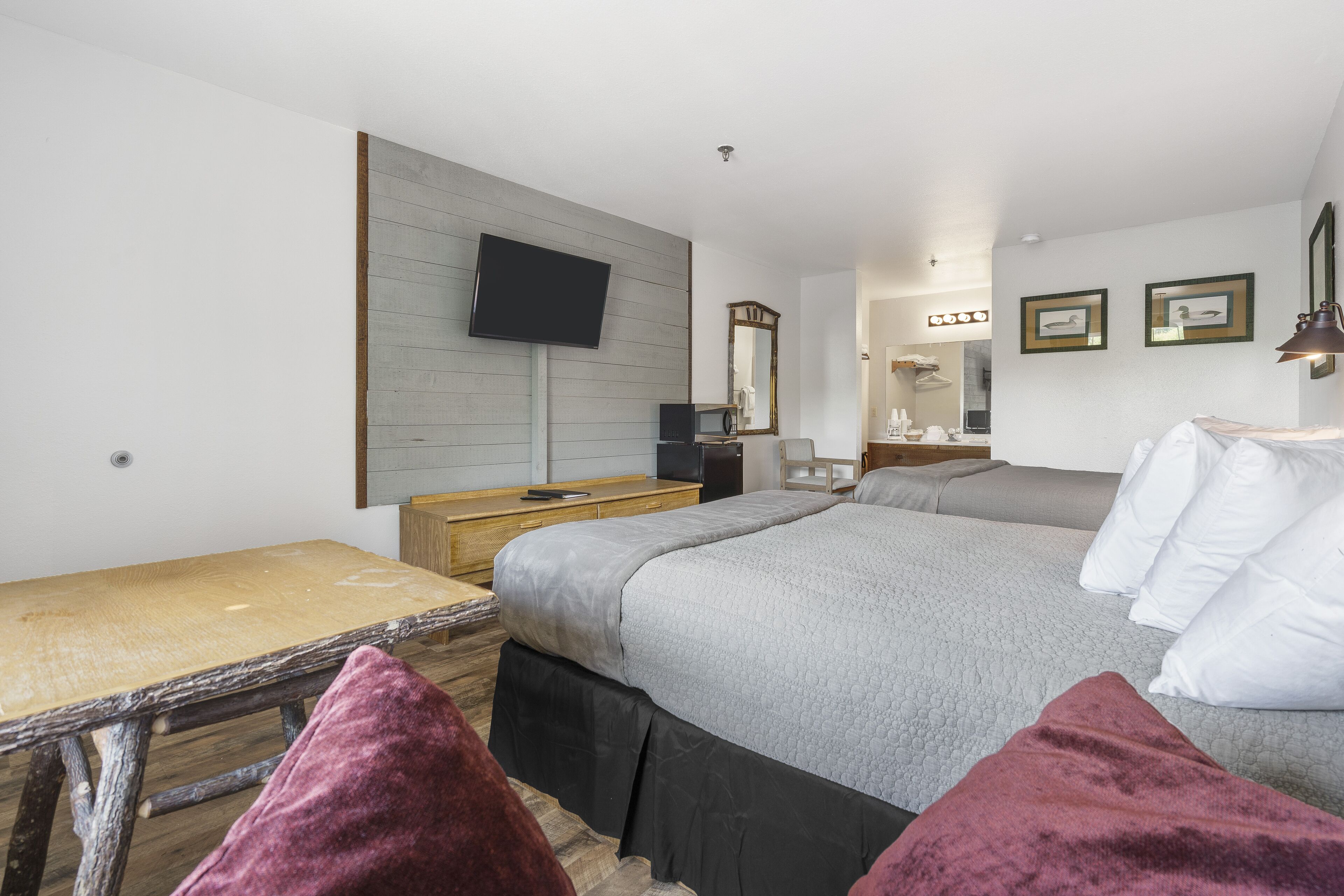 A bedroom with a wooden desk, a bed with a grey quilt, a television mounted on the wall, and a mirror above the dresser.