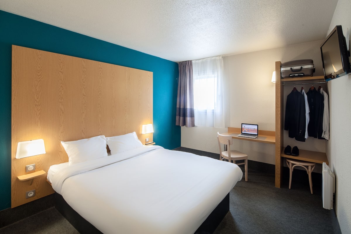 Une chambre d’hôtel avec un grand lit, des lampes de chevet, un bureau avec un ordinateur portable, une chaise, un porte-manteau et un téléviseur.
