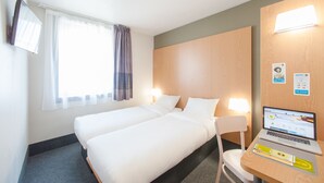 Quarto de hotel com duas camas, uma escrivaninha com um laptop, uma cadeira, uma janela com cortinas e uma televisão na parede.