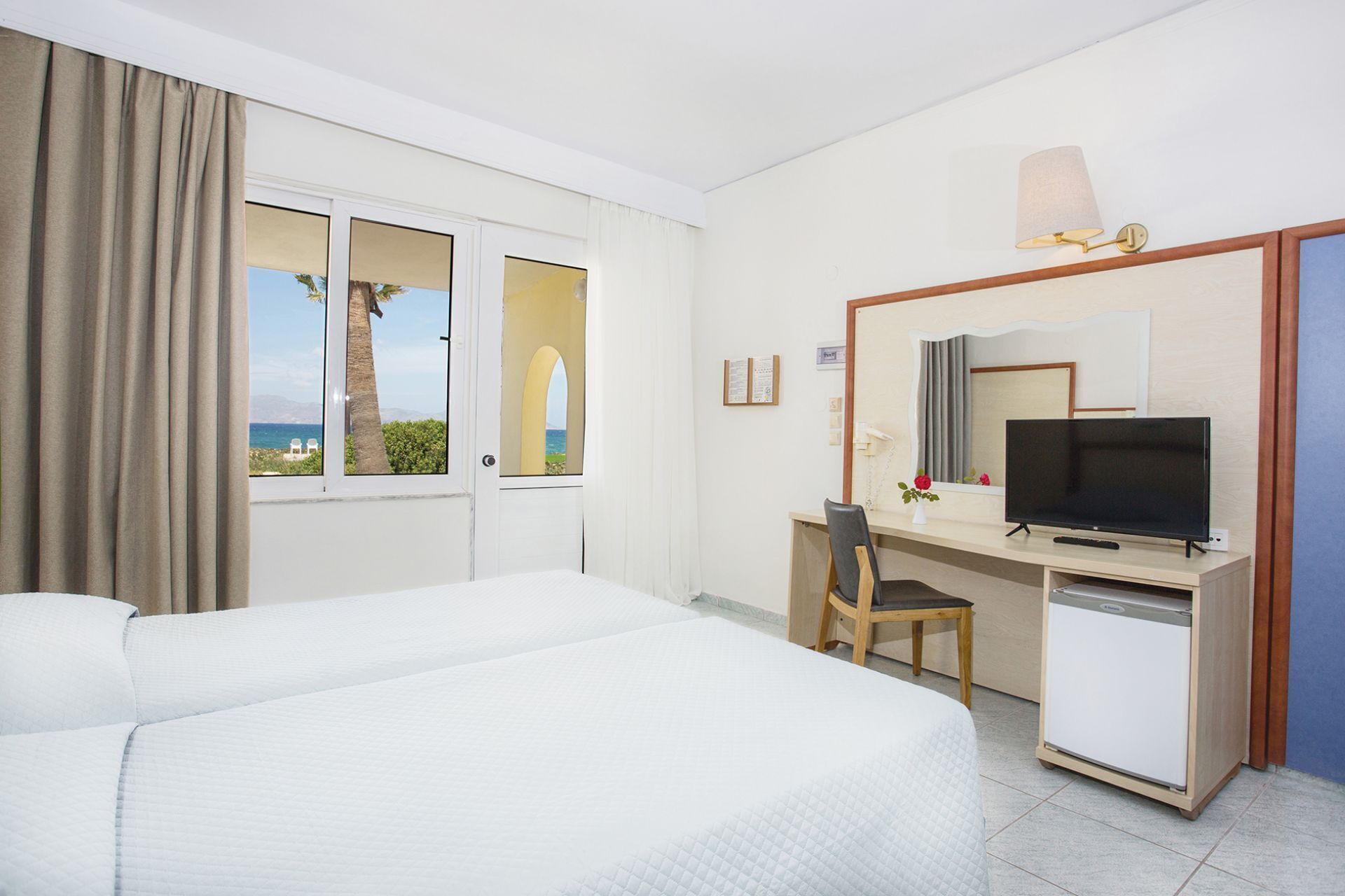 Habitación de hotel con cama, escritorio con televisor, refrigerador pequeño y ventana con vista al mar.