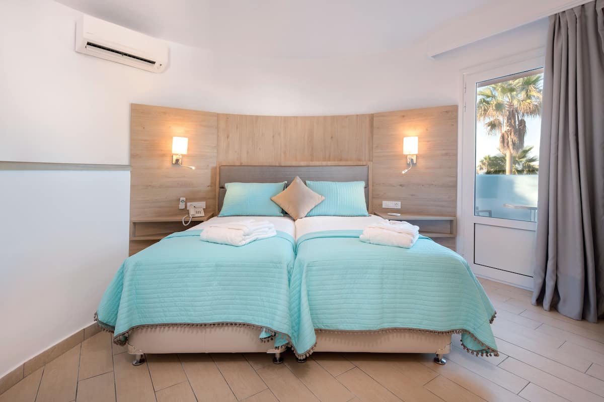Un lit double recouvert d’une couvre-lit bleu clair, deux tables de chevet avec des lampes, une tête de lit en bois et une vue sur des palmiers à travers une fenêtre ornée de rideaux.