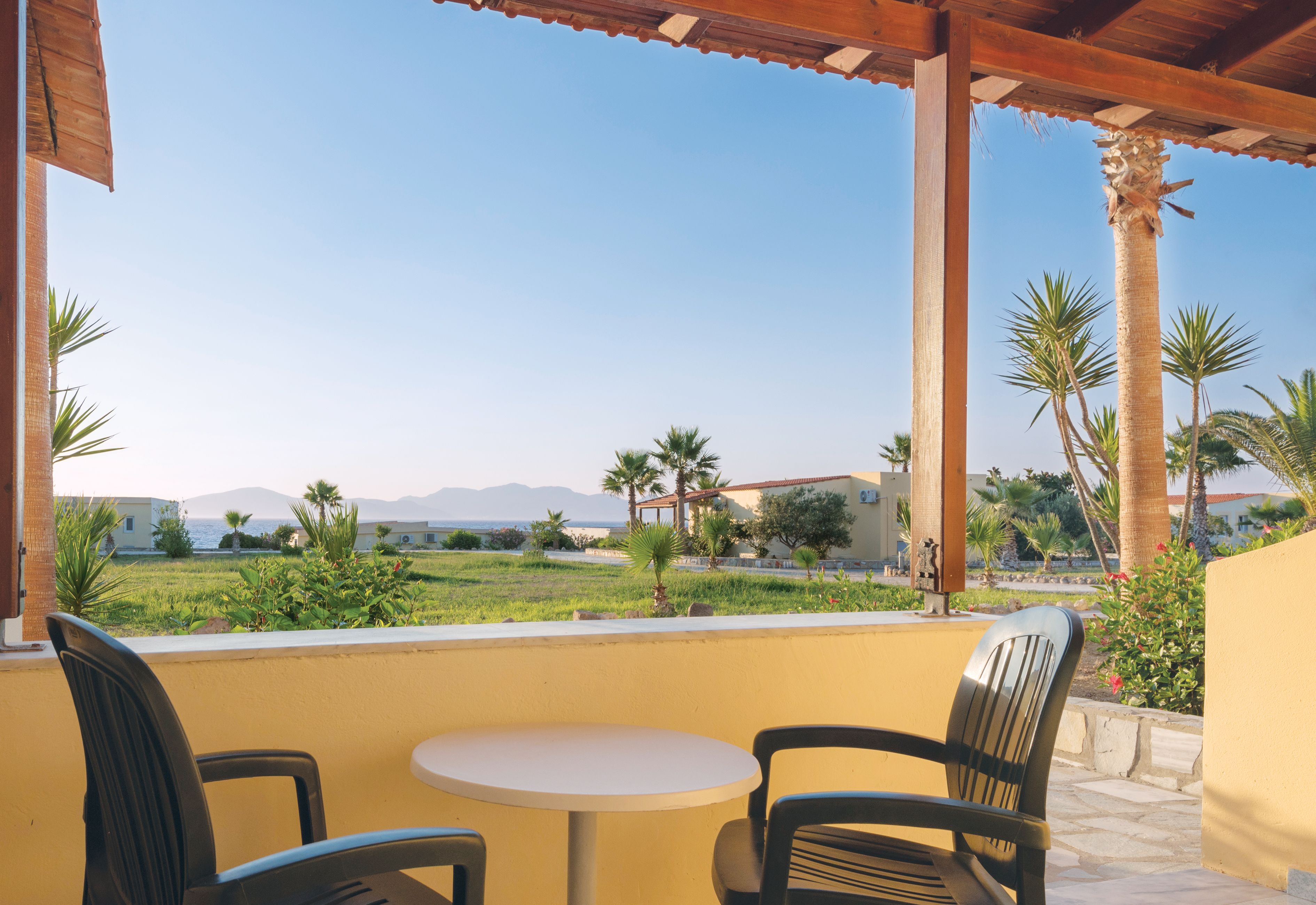 Un patio exterior con dos sillas y una mesa, con vistas a una zona ajardinada con palmeras y una lejana vista de edificios y montañas.