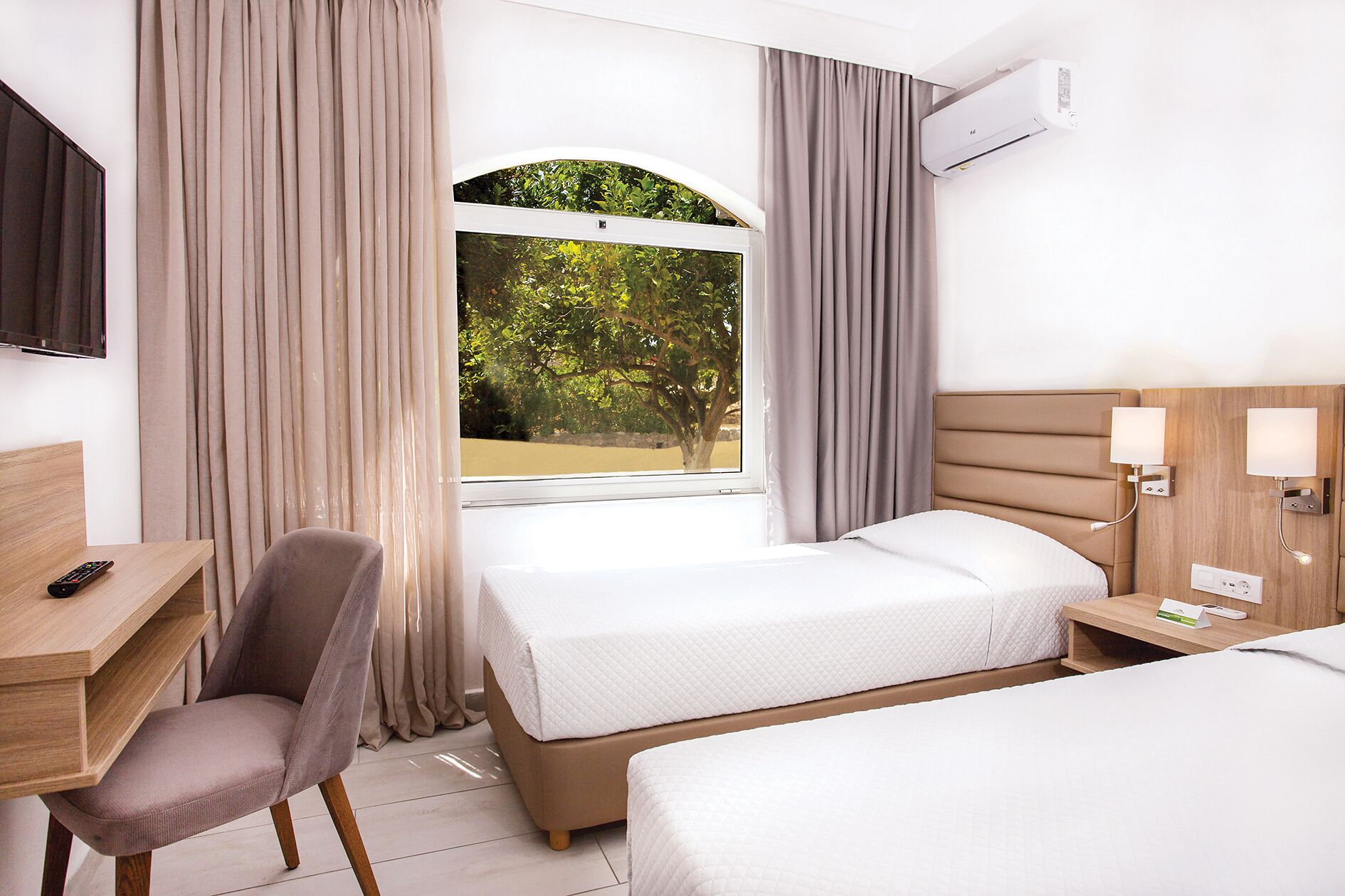 Quarto de hotel com duas camas, uma mesa, uma cadeira, televisão, ar condicionado e janela com cortinas.