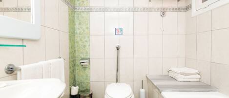 Un baño con inodoro, lavabo y bañera. Hay toallas apiladas en la pared y un cubo de basura.