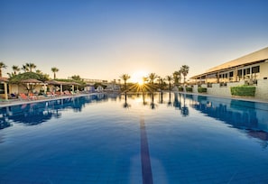 A swimming pool with lounge chairs and umbrellas, surrounded by palm trees and a building with large windows.