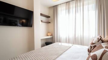 Una camera d'albergo moderna con una TV a schermo piatto montata a parete, un letto con lenzuola decorate, un comodino con un vaso e tende leggere alla finestra.