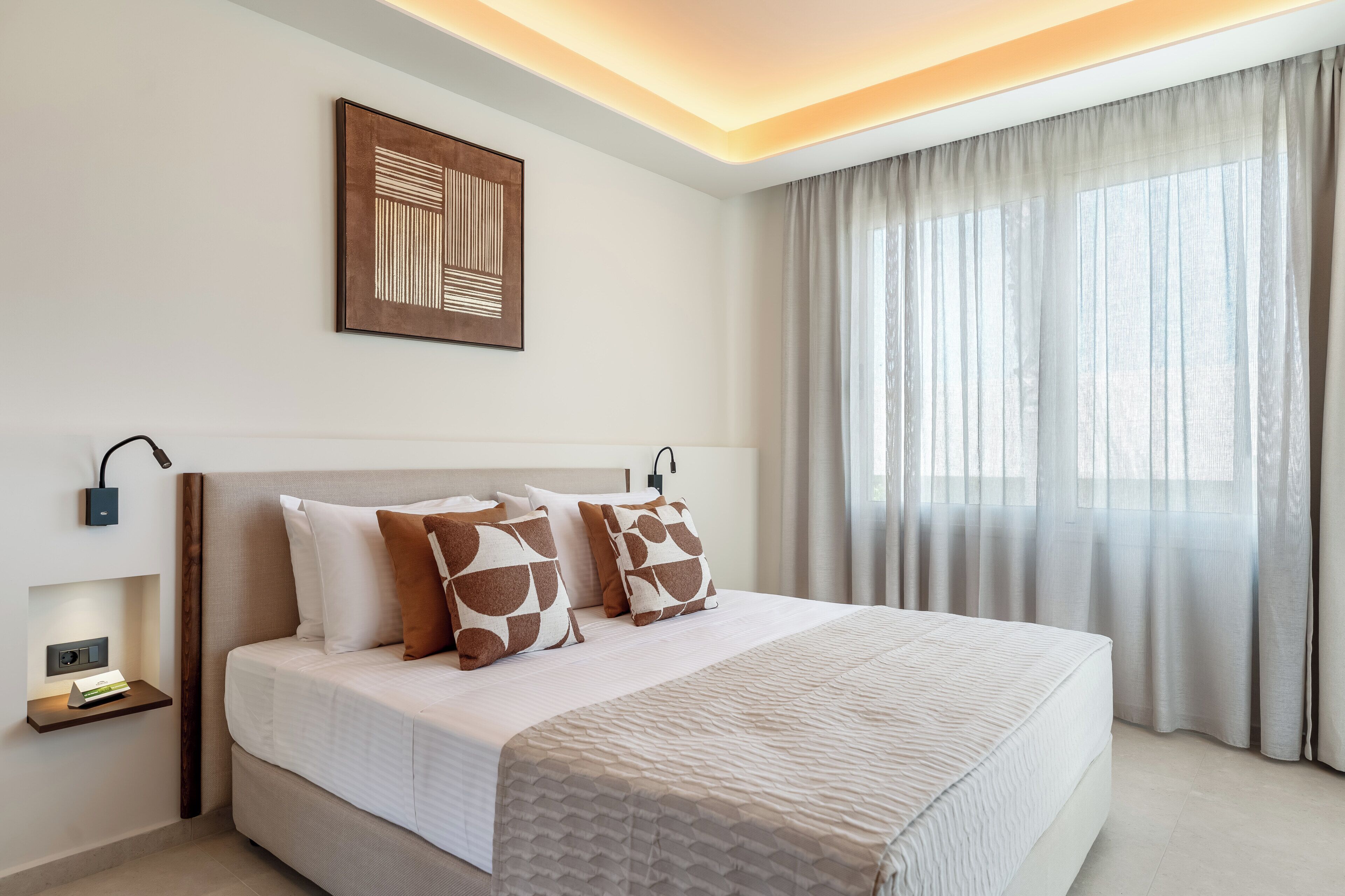 Una habitación de hotel moderna con una cama grande, una mesita de noche con lámpara, una obra de arte colgada en la pared y un amplio ventanal con cortinas.