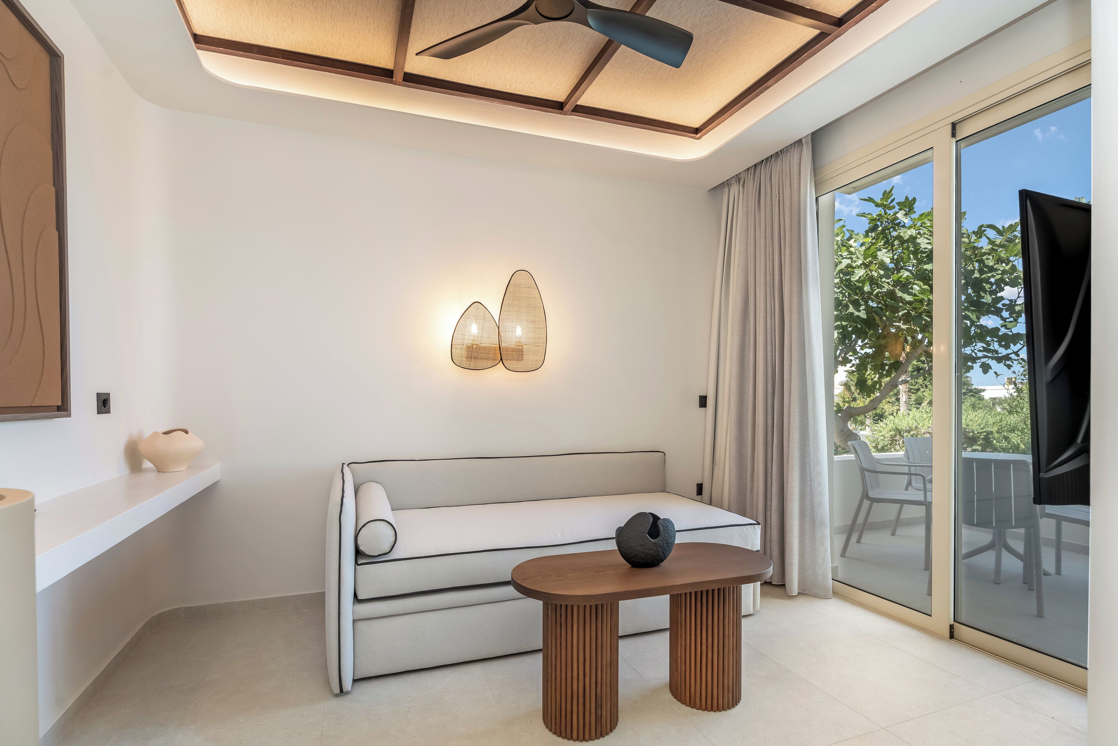 Una sala de estar moderna con un sofá, una mesa, un ventilador de techo y un amplio ventanal corredera que da a un área exterior.