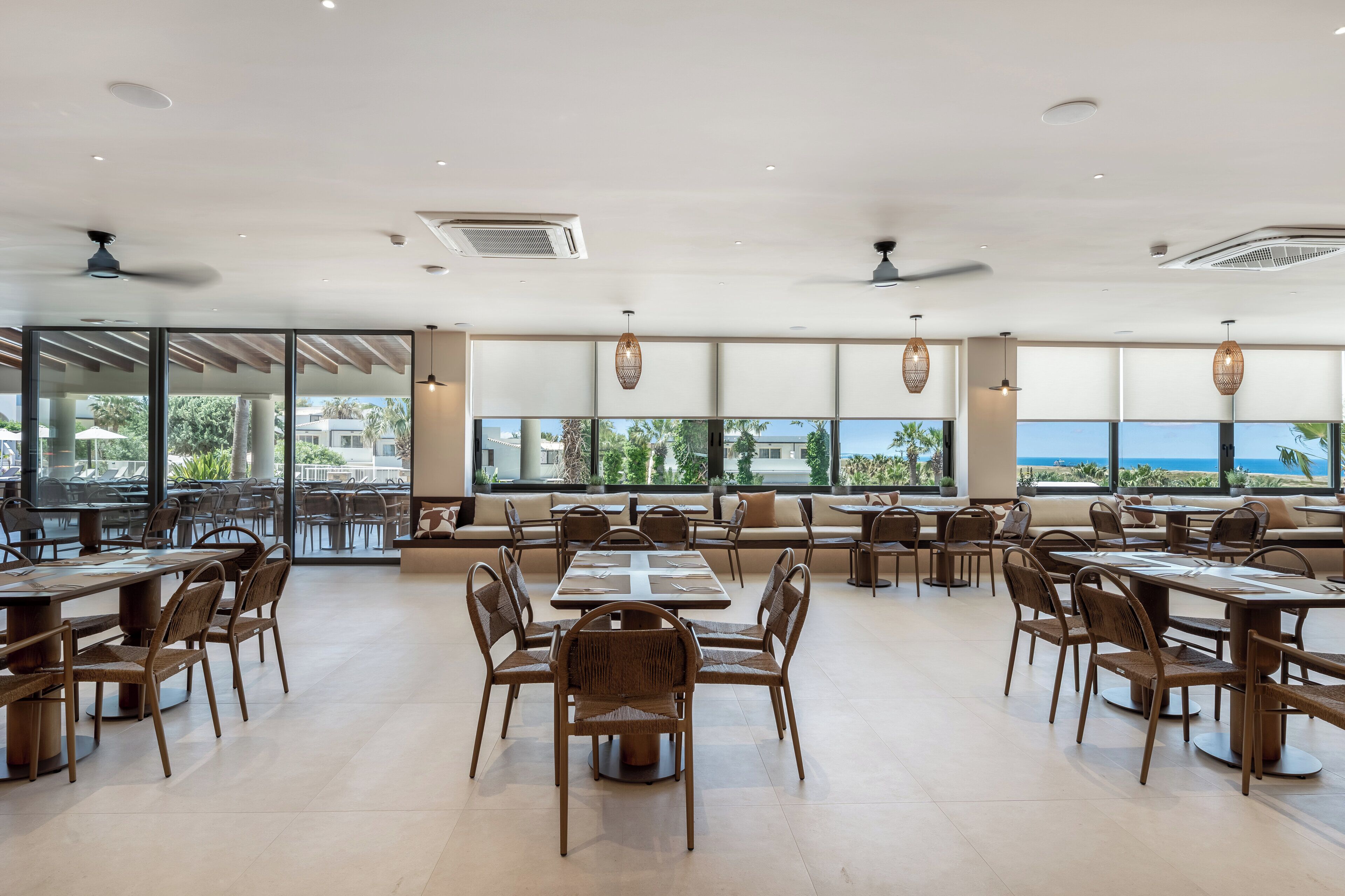 Uma ampla área de jantar com várias mesas e cadeiras, grandes janelas com vista para o mar e ventiladores de teto.