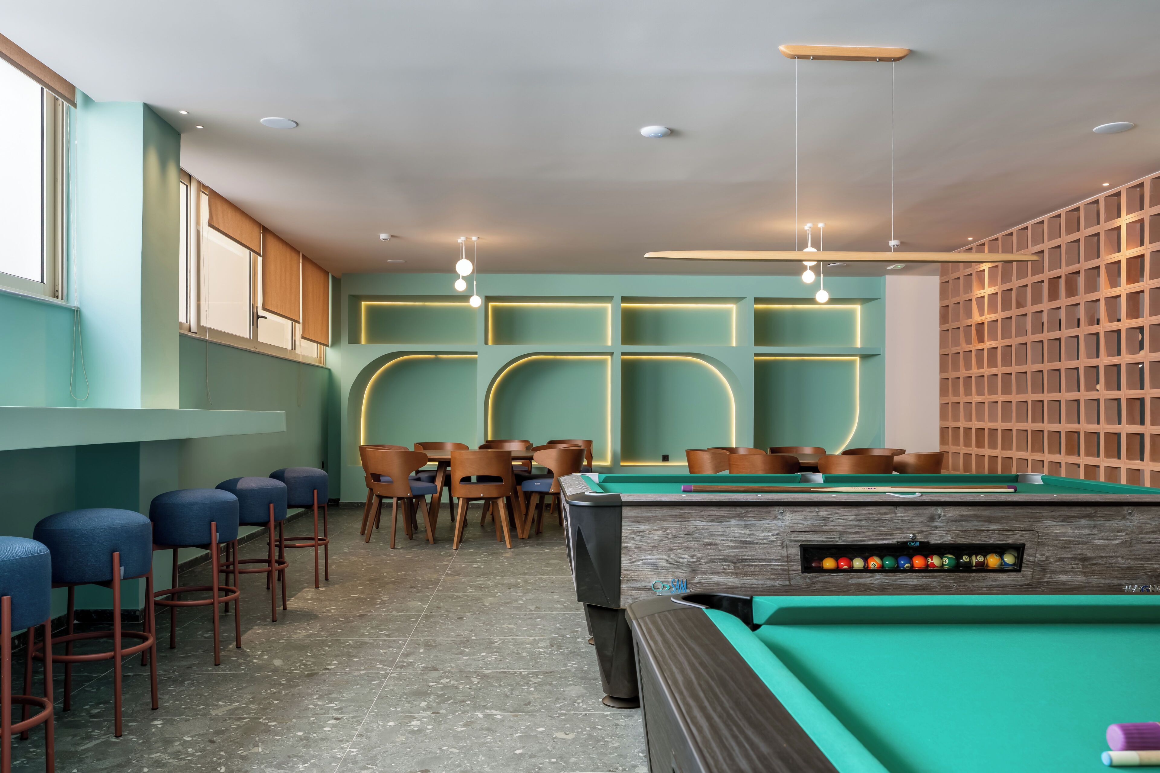 Mesa de bilhar com cadeiras e área de bar em um ambiente com paredes verde-água e prateleiras de madeira.