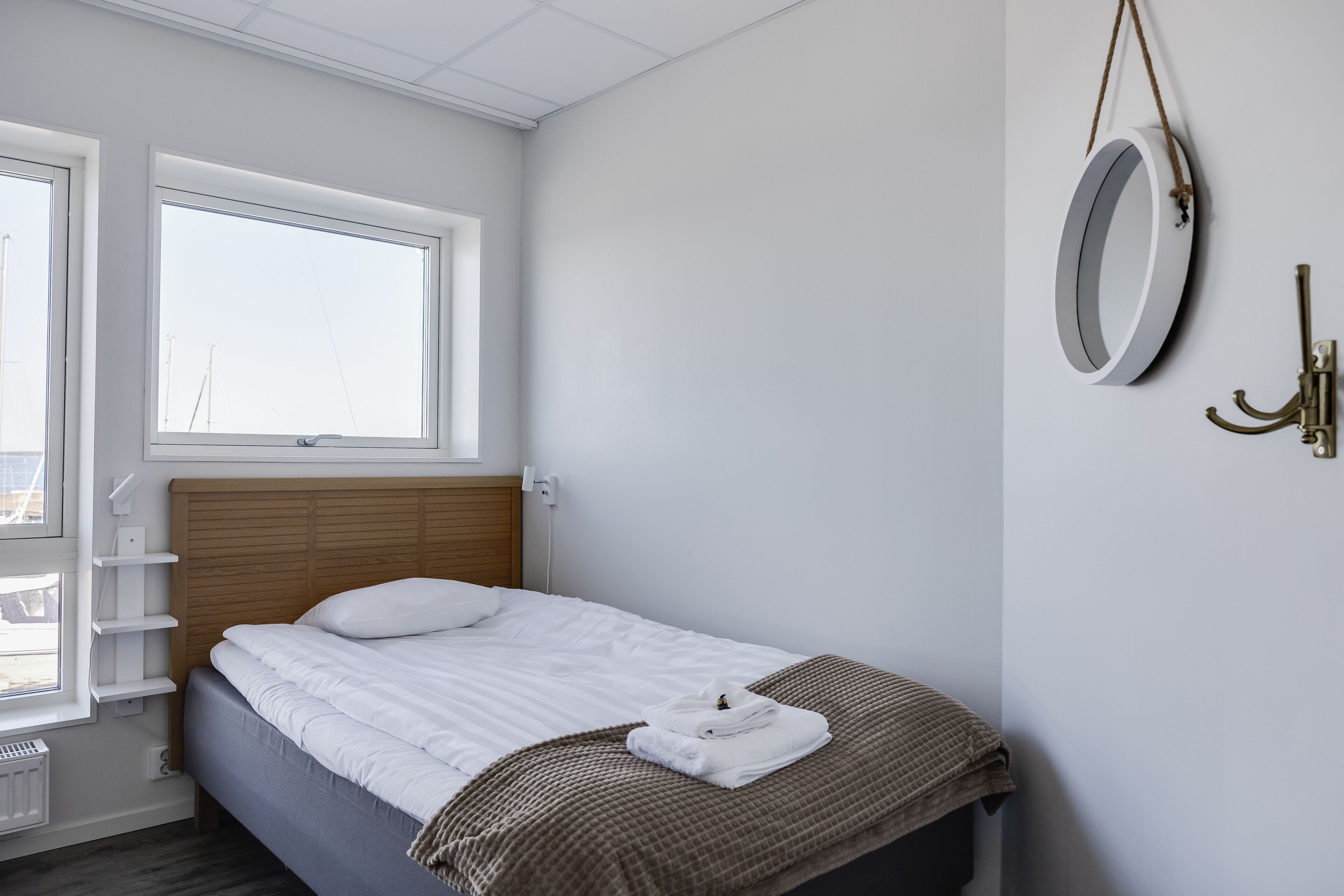 Ein ordentlich gemachtes Bett mit weißen Leinen, einer braun karierten Decke und einem zusammengeklappten Handtuch auf der Seite. Ein Fenster mit Blick nach draußen und ein an der Wand befestigter Spiegel mit Messingrahmen.