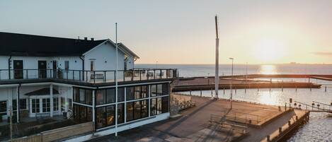 Een gebouw aan het water met een glazen gevel, een overdekt terras en uitzicht op de ondergaande zon.
