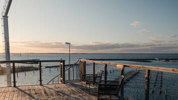 A wooden deck with metal railings overlooking a body of water, a distant bridge, and a clear sky.