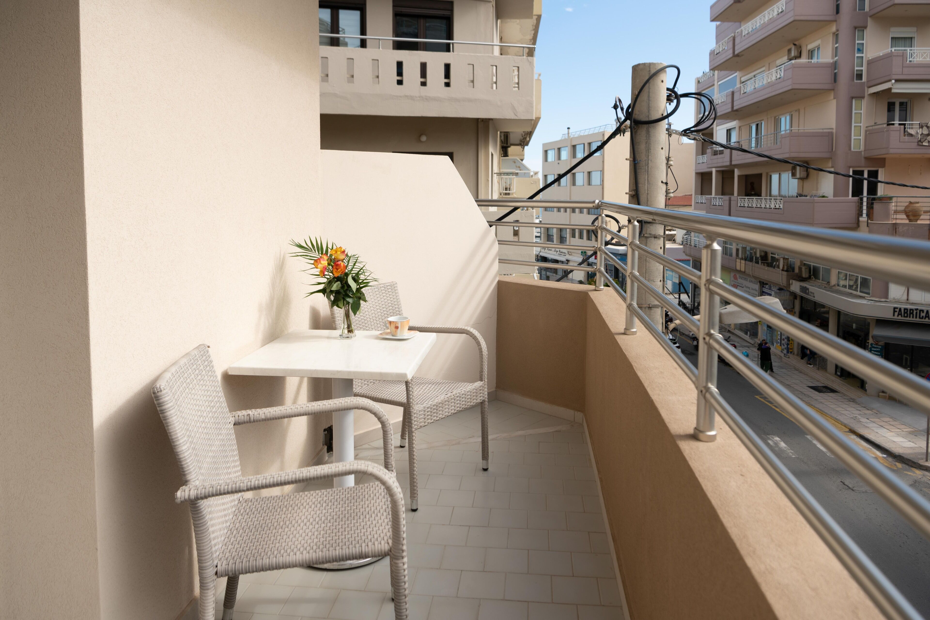 A balcony with a white table and wicker chairs, a vase with flowers, and a view of the street and buildings.