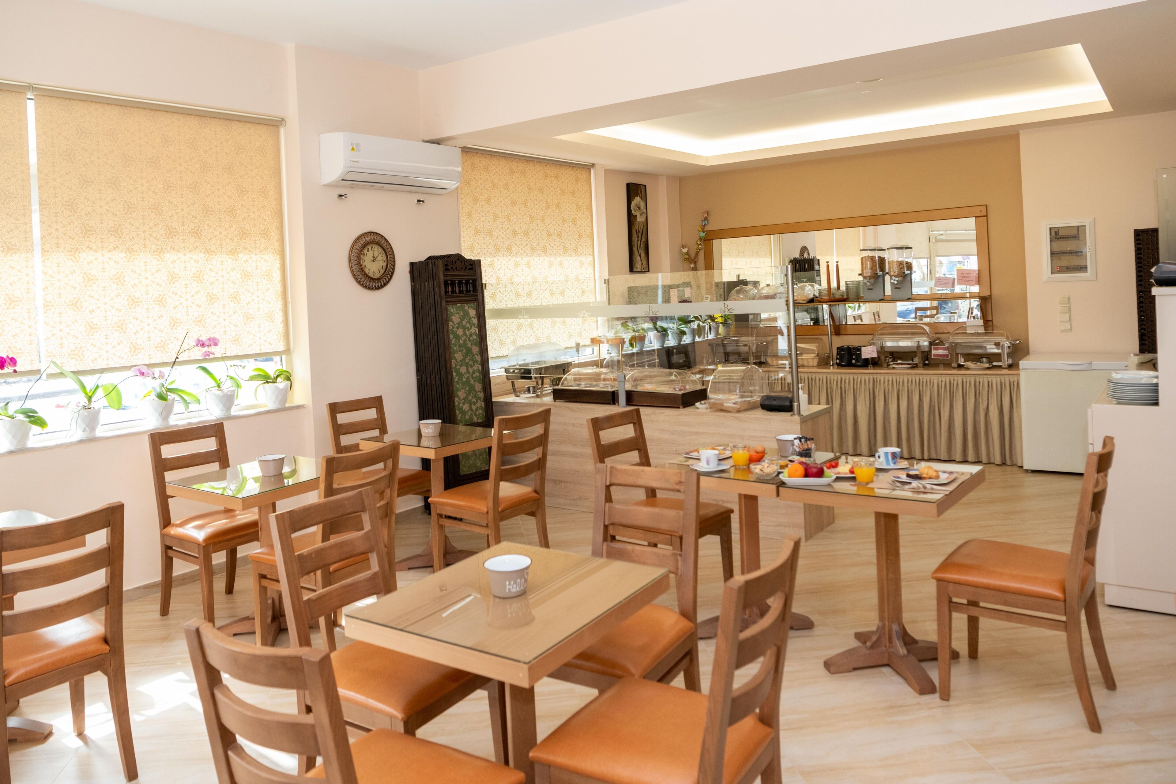 A dining area with multiple tables and chairs, a kitchen in the background, and a window with potted plants.