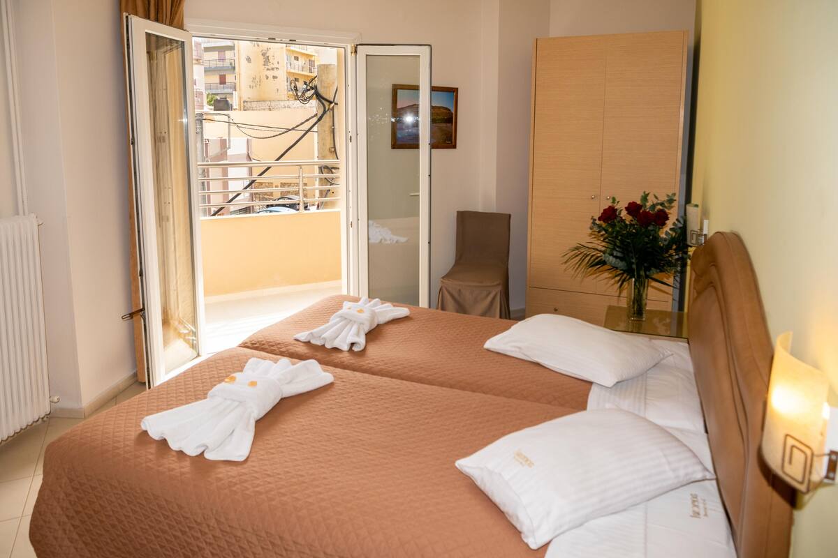 Une chambre d’hôtel avec deux lits, un balcon, une armoire en bois et un vase rempli de fleurs.