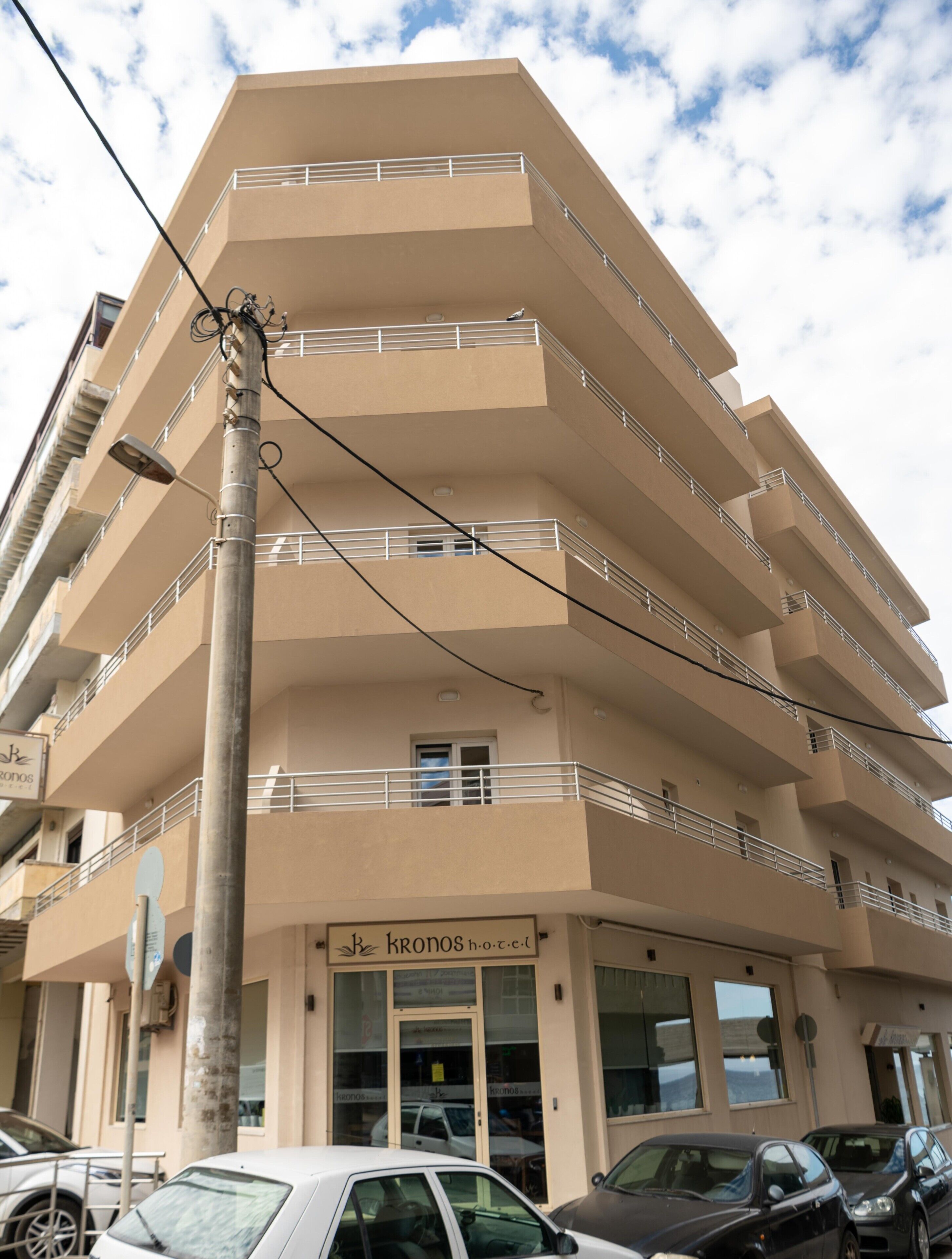 Un edificio de varios pisos con balcones, un letrero que dice "KRONOS hotel" y coches estacionados en la parte delantera.