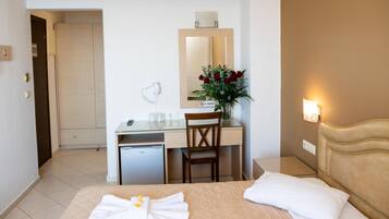 A hotel room with a bed, a desk, a chair, a small fridge, a mirror, a vase with flowers, and a wall-mounted light.