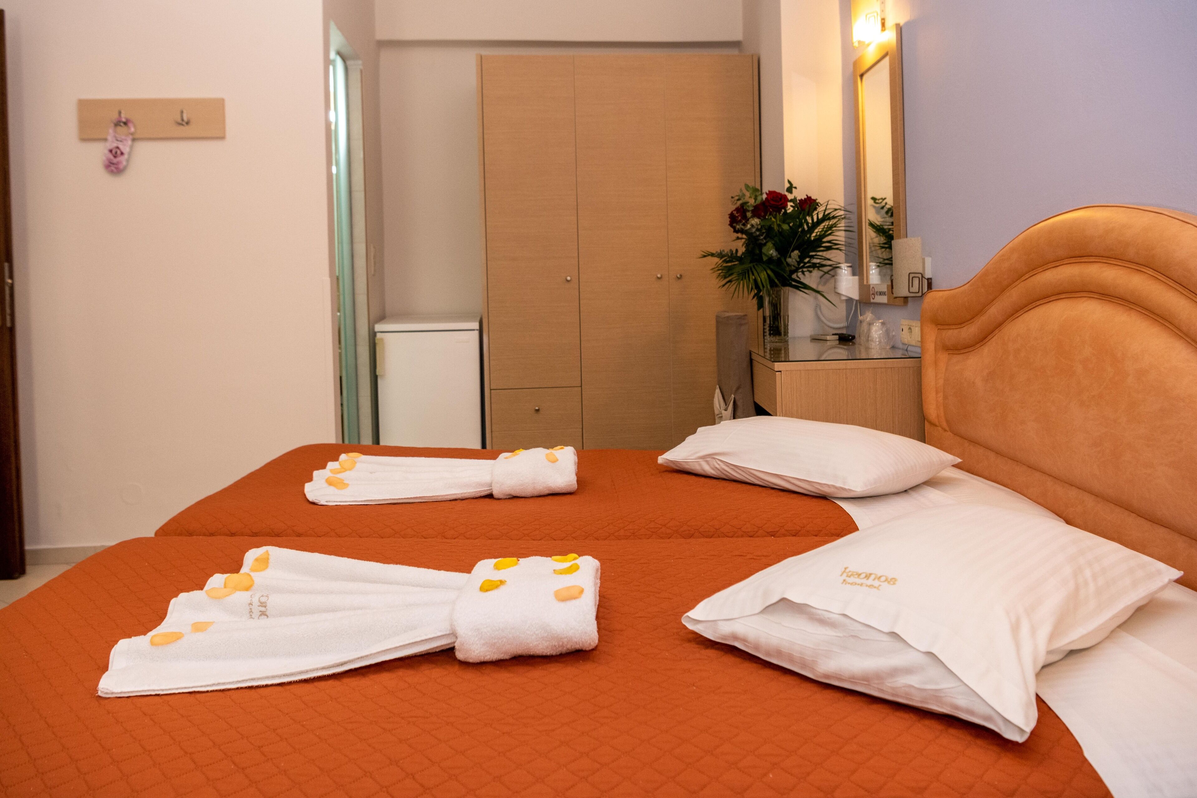 Habitación de hotel con dos camas, cada una con ropa de cama naranja y toallas blancas con detalles en naranja. Se observa un armario de madera, una mesita de noche con un jarrón de flores y un espejo en la pared.