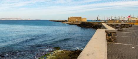 Vista costera con un edificio histórico de piedra, un camino pavimentado y un puerto deportivo al fondo.