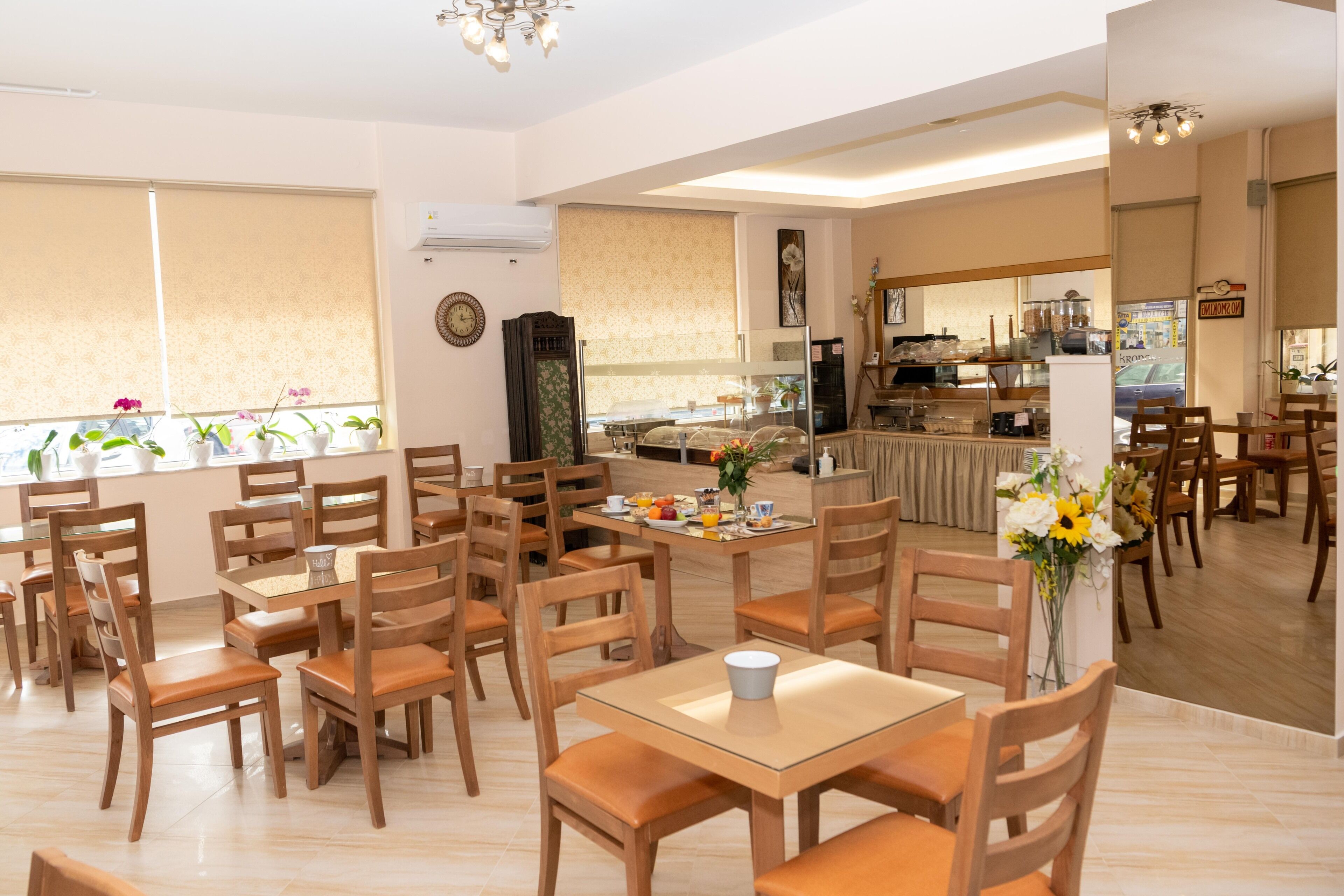A spacious dining area with wooden tables and chairs, a kitchen in the background, and a large window with blinds.