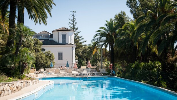 A swimming pool with lounge chairs and umbrellas, surrounded by palm trees and a building with a tiled roof. - Residence RTA Villa Marina (Imperia)