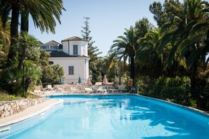 Une piscine entourée de palmiers et d’un bâtiment à toit de tuiles, avec des chaises longues et des parasols à proximité.