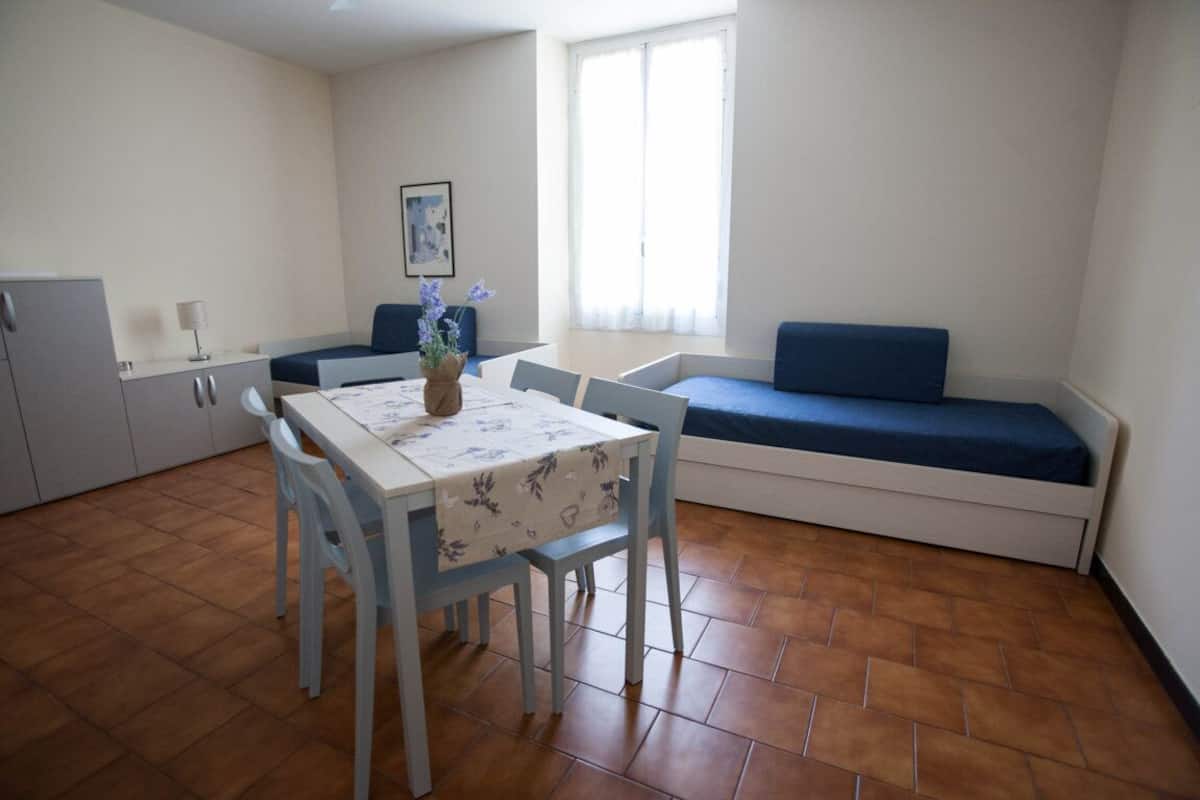 Una habitación pequeña y luminosa con una mesa de comedor, sillas, un sofá con un cojín azul y una ventana que deja entrar luz natural.