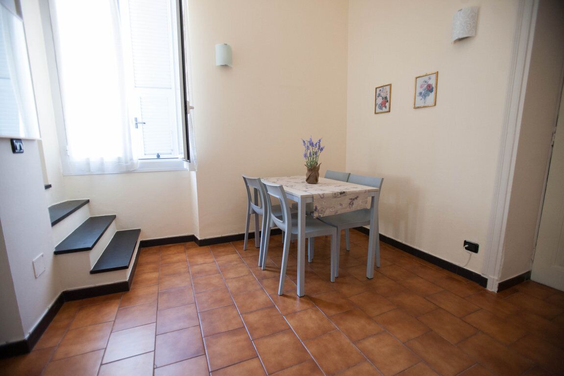 A dining area with a table, chairs, and a vase of flowers. There are steps leading to a door and a window with blinds.