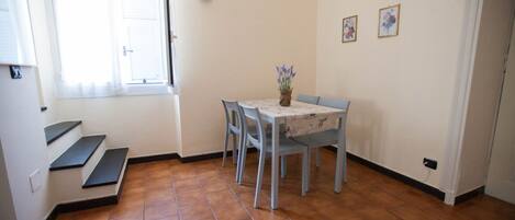 Un comedor con una mesa, sillas y un jarrón de flores. Se ven escalones que llevan a una puerta y una ventana con persianas.