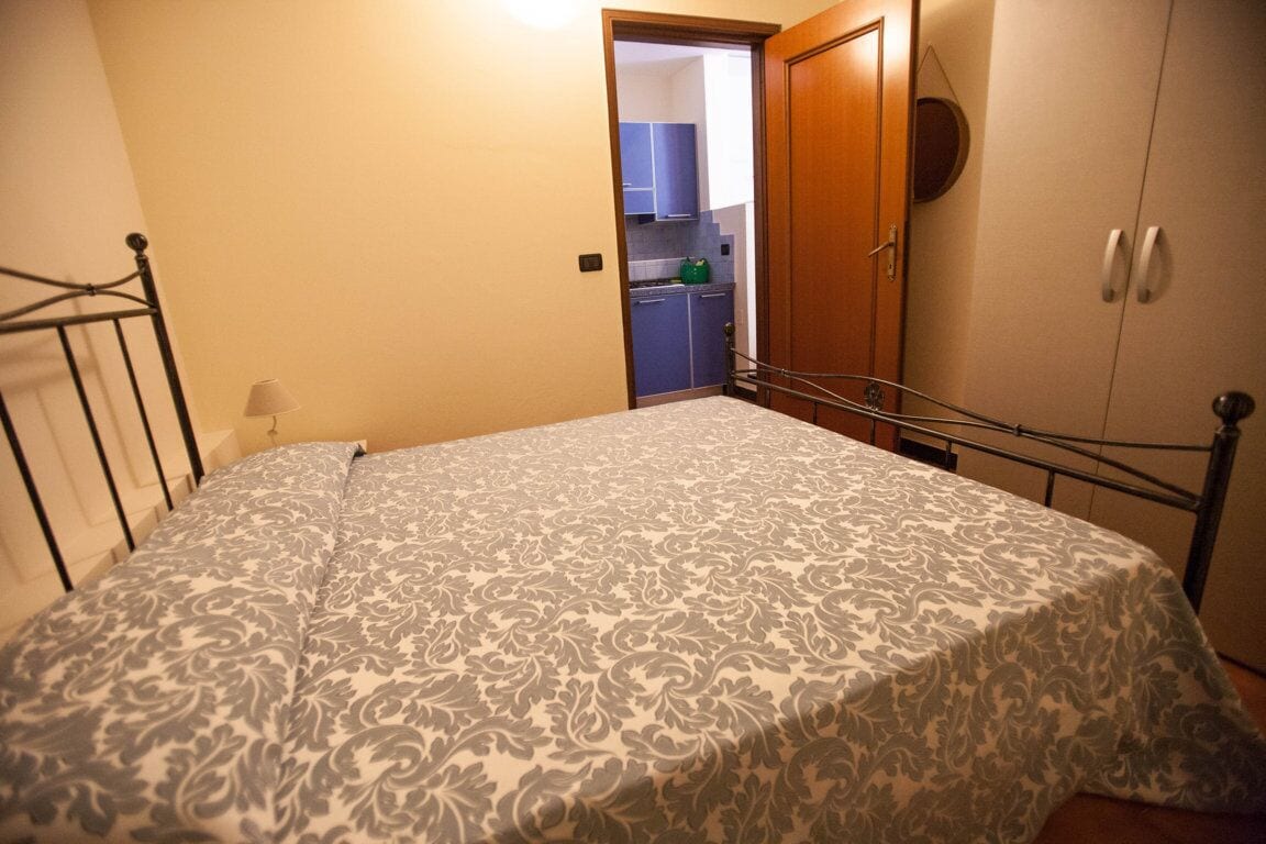 A bedroom with a bed, a wardrobe, and a door leading to a kitchen.