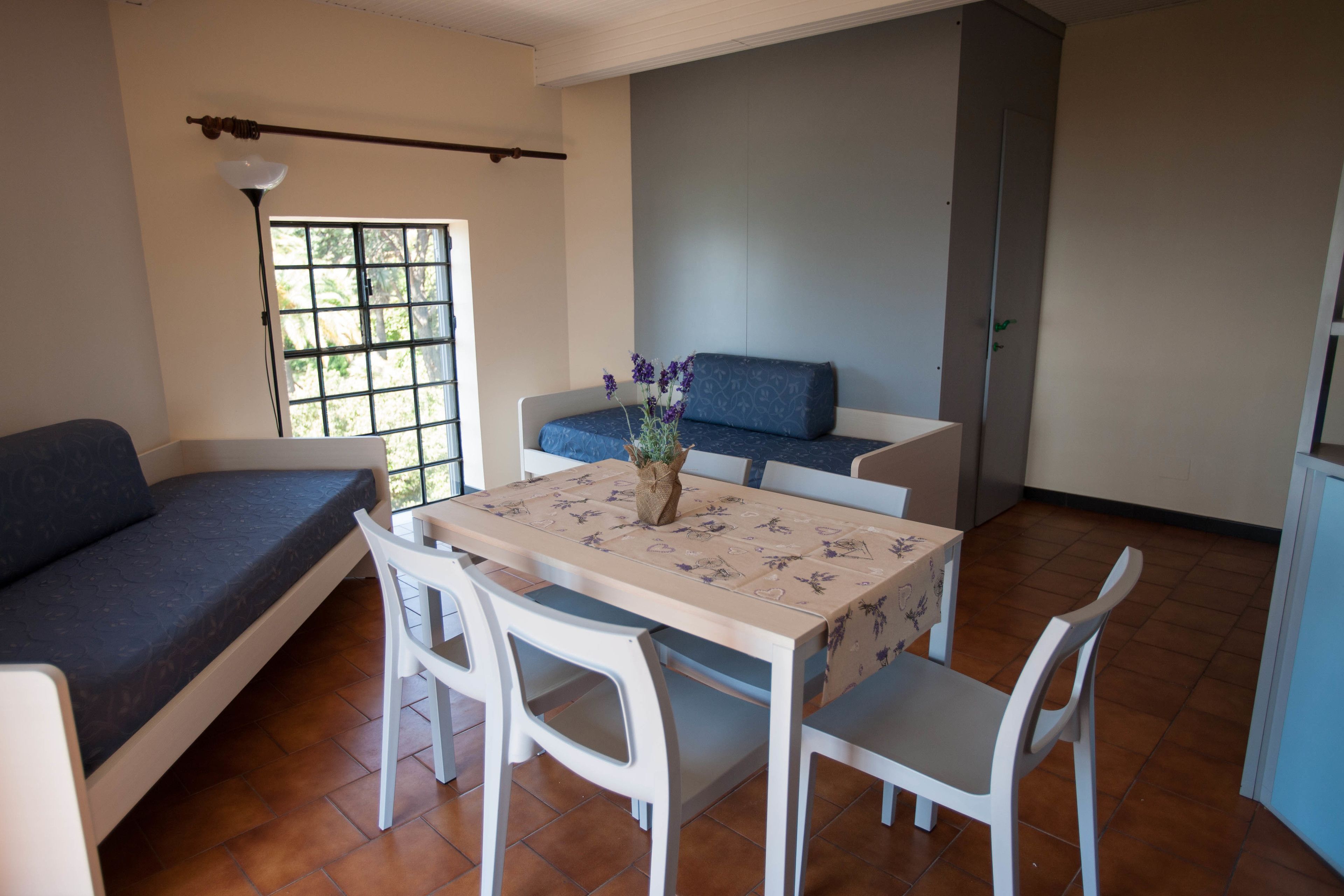 Un espacio habitable compacto con sofá, una mesa de comedor con sillas, una pequeña cocina y un ventanal que deja entrar luz natural.