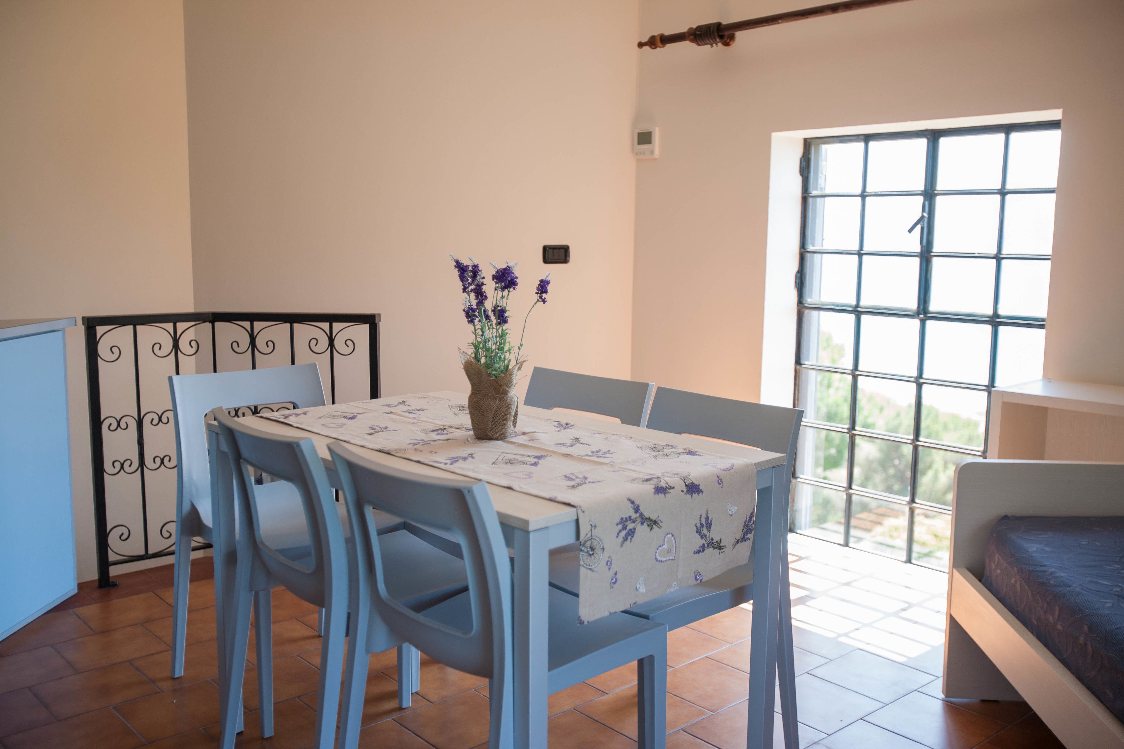 A dining area with a table set for two, a vase with flowers, and a window with a view of greenery.