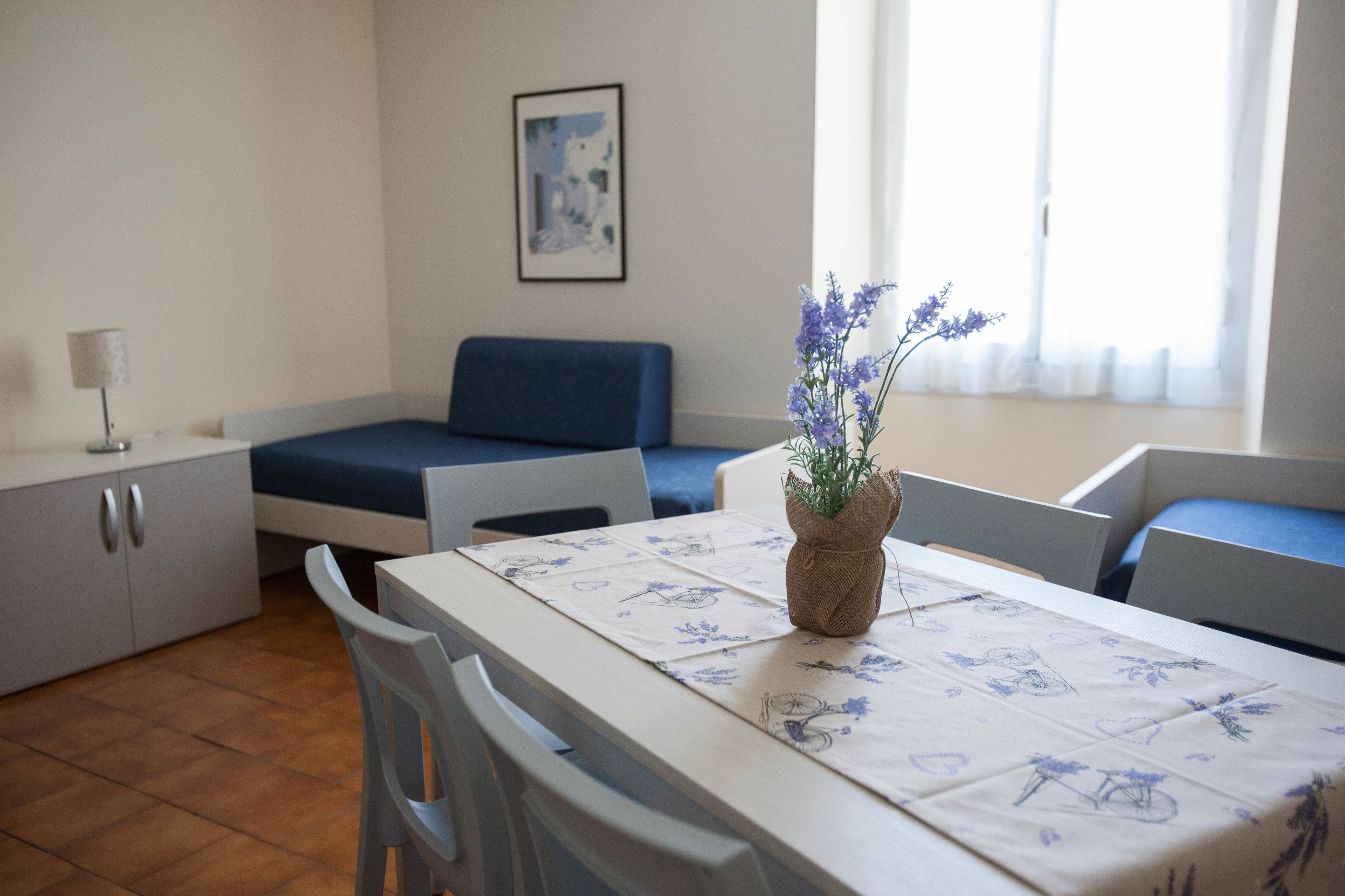 Une pièce comprenant une table à manger, des chaises, un canapé, un petit meuble, une lampe et une fenêtre avec des rideaux.