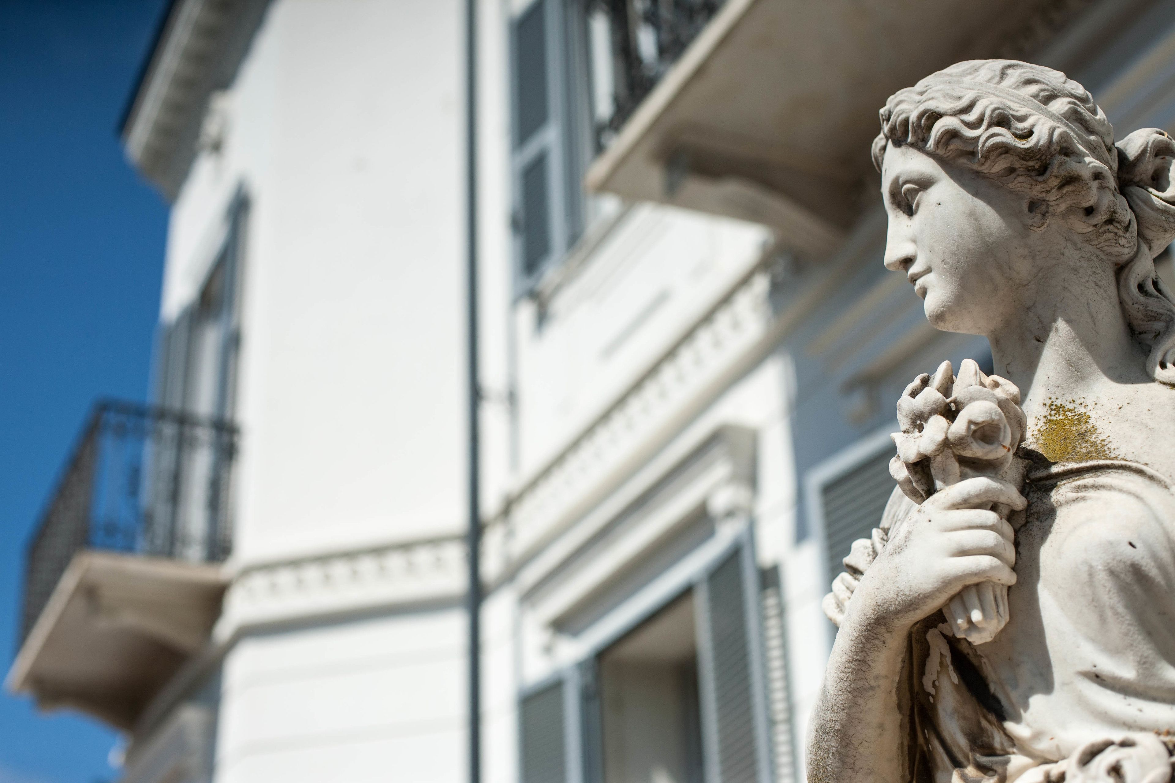 Una statua raffigura una figura con un grappolo d'uva, posta di fronte a un edificio dalle caratteristiche architettoniche classiche.
