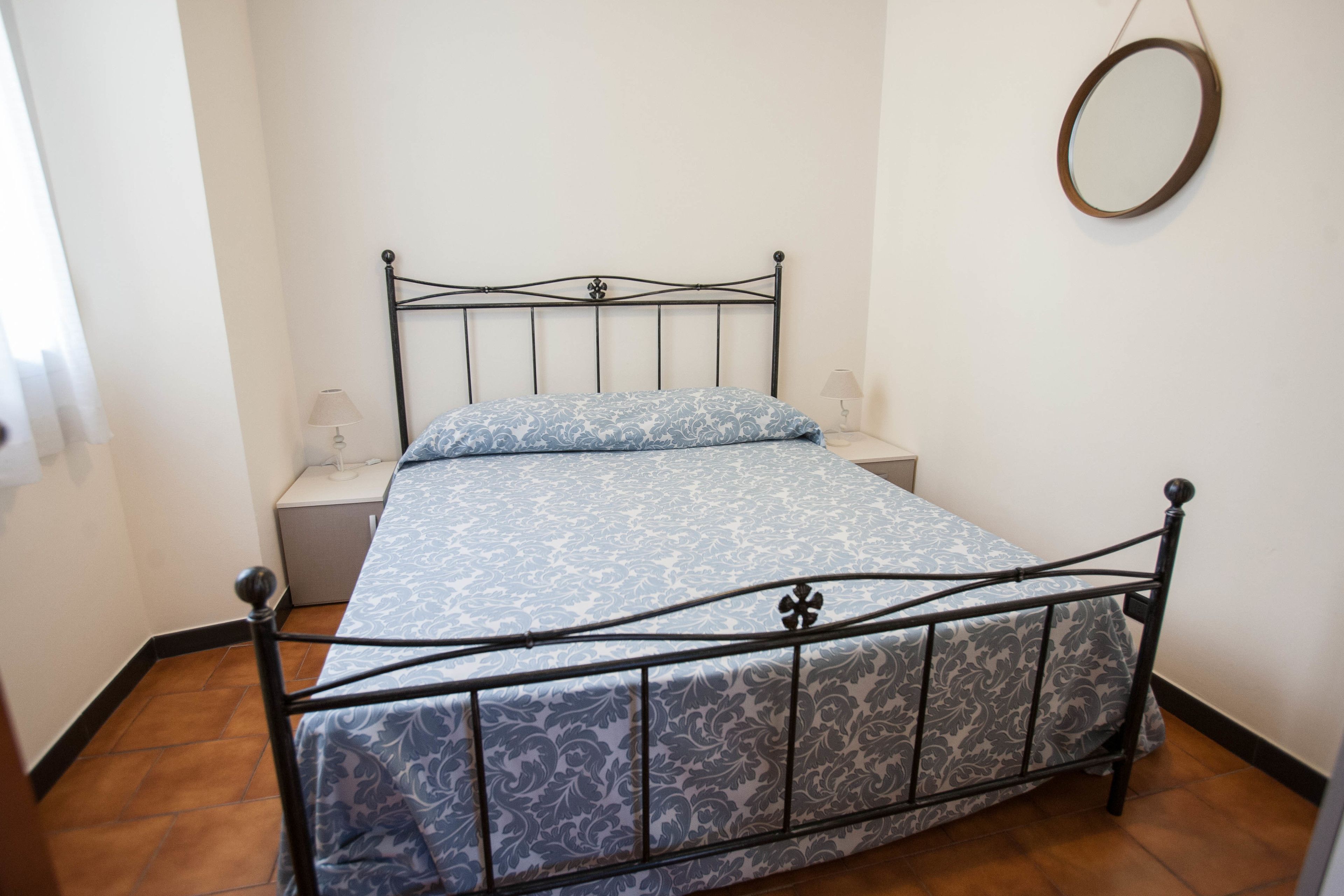 A single bed with a metal frame and patterned bedding, a bedside table with a lamp, and a round mirror on the wall.