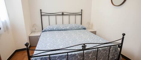 A single bed with a metal frame and patterned bedding, a bedside table with a lamp, and a round mirror on the wall.