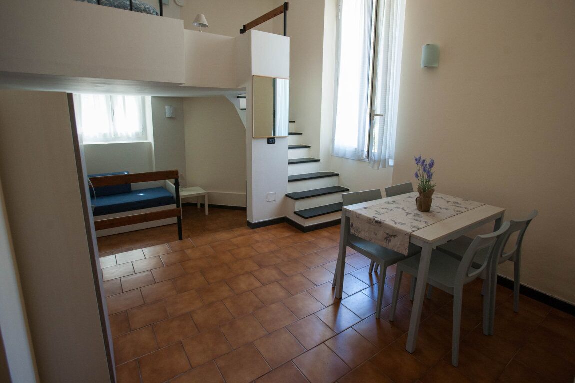 Una habitación con literas, una escalera, una mesa de comedor con sillas y un florero con flores.