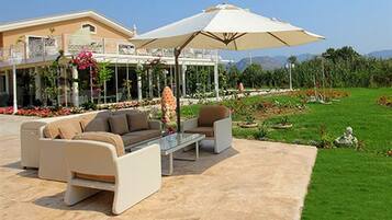 Un espace extérieur aménagé avec un canapé, des fauteuils et une table, entouré d’un jardin fleuri et abrité par un grand parasol.