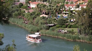Un barco de paseo fluvial con cubierta techada y pasajeros, rodeado de exuberante vegetación y edificios residenciales con techos rojos.